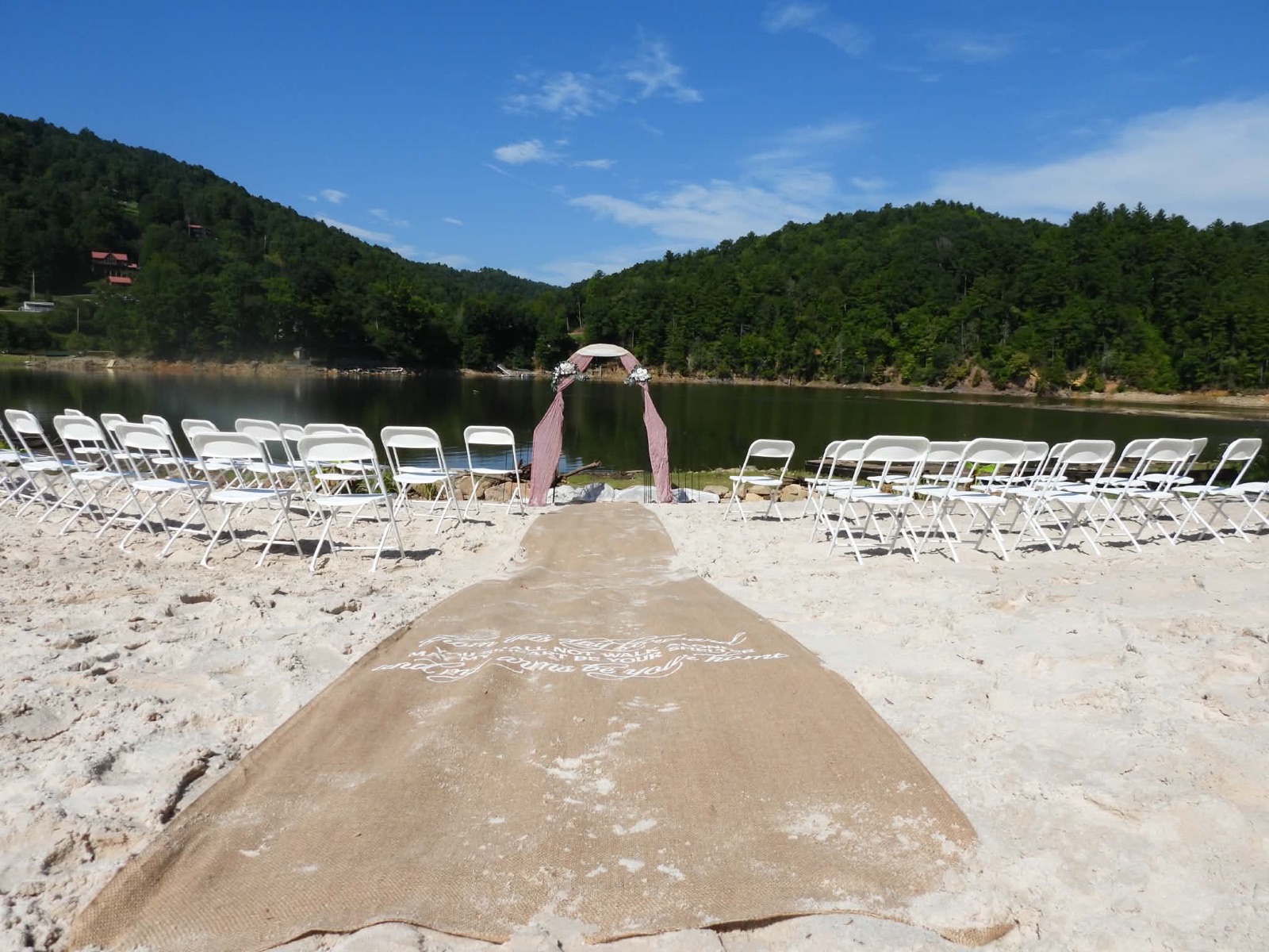 Beach wedding ceremony set up on the private white-sand beach — burlap aisle runner leading to a draped arch at the water's edge